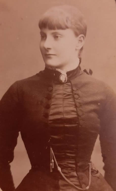 Sepia coloured image of Norah Guthrie (née Power), one of five children of Martha and William Power, and the mother of Tyrone and Susan Margaret Guthrie. Norah is wearing a late Victorian or early Edwardian dark dress with buttoned over jacket. She is wearing a cameo brooch at her neck, and from the viewers perspective, a thick gold chain, possibly a key or watch chain, between her right waist and a button on left side of her jacket. Norah’s hair is worn with a fringe, with the suggestion of a ponytail at her back. She is looking facing the camera but looking away from the viewer.